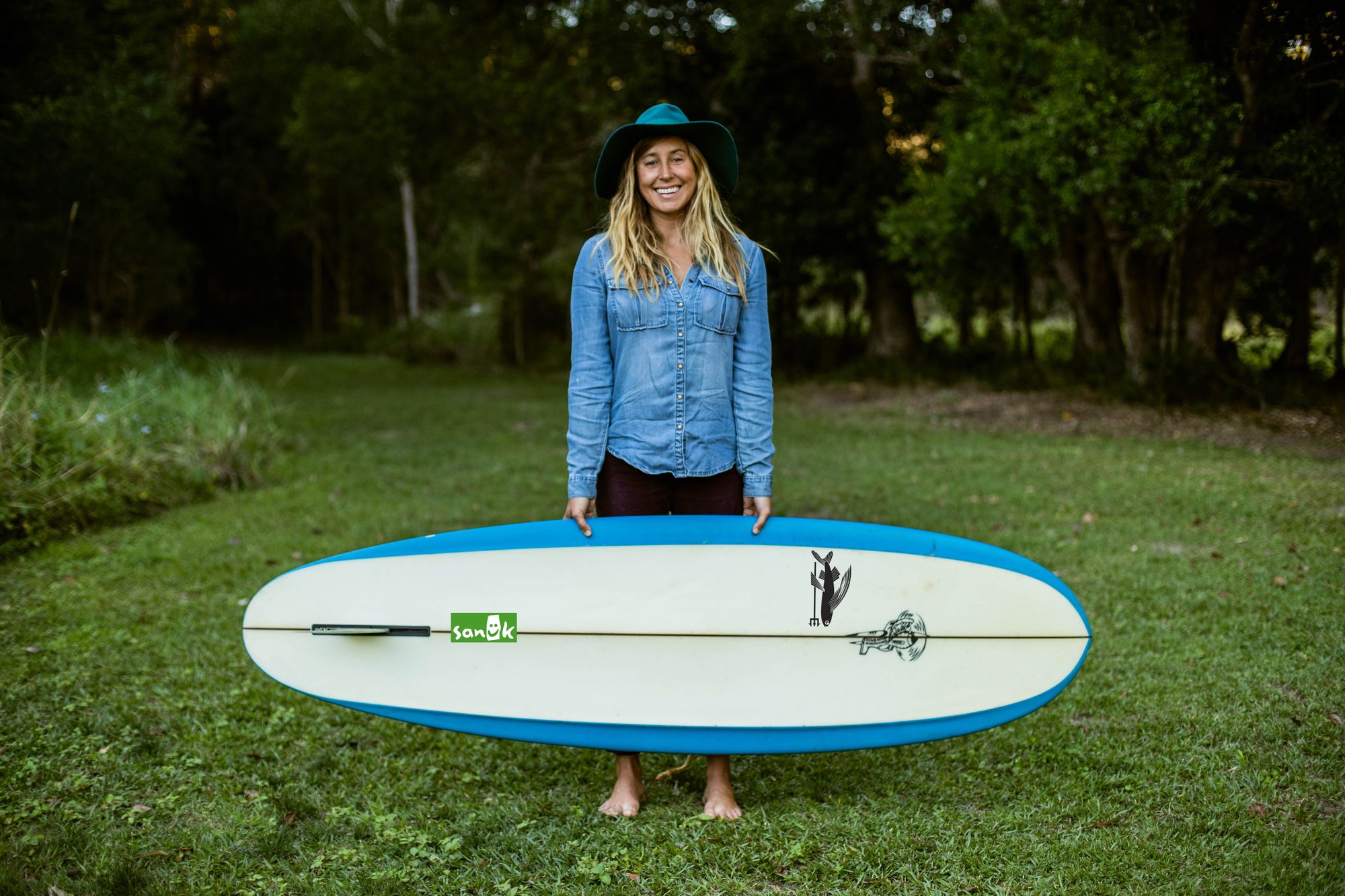 Lauren Hill stands in a plain of grass, with trees behind her, holding her surfboard and smiling.