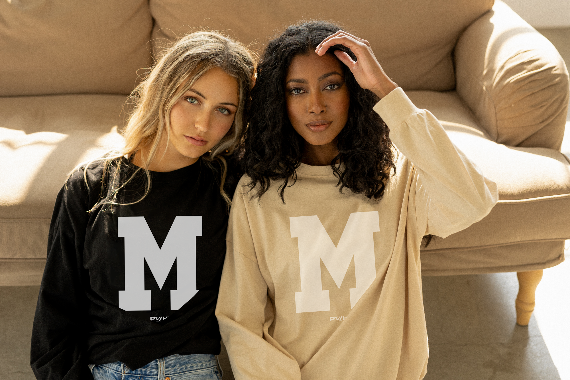 Photo of two women sitting on the floor next to a beige couch, each wearing a long-sleeve t-shirt with a large white M logo.